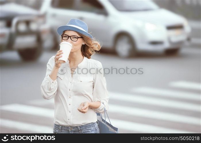 Happy young fashionable student drinking hot take away coffee and crossing a road against urban background.