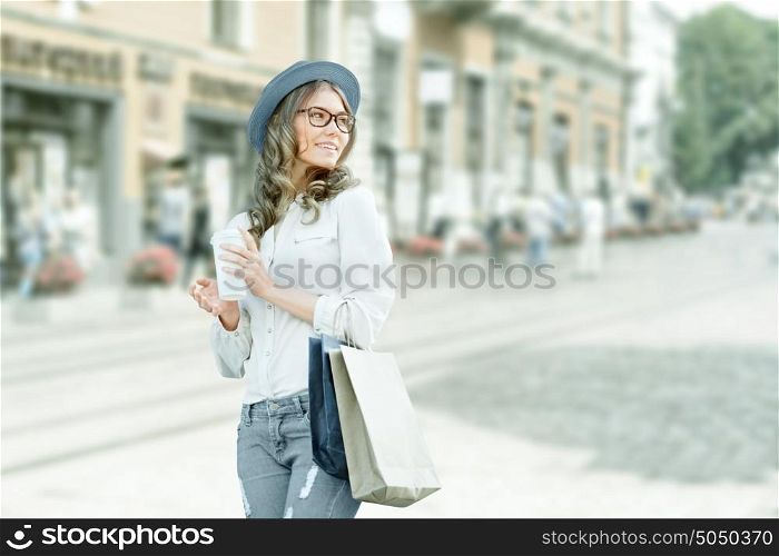 Happy young fashionable woman taking a coffee break after shopping, smiling with a coffee-to-go in her hands against urban city background.