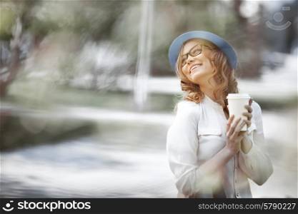 Happy young fashionable woman taking a coffee break after shopping, smiling with a coffee-to-go in her hands in a summer park.