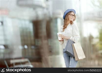 Happy young fashionable woman with bags having a coffee break after shopping and holding take away coffee against urban background.