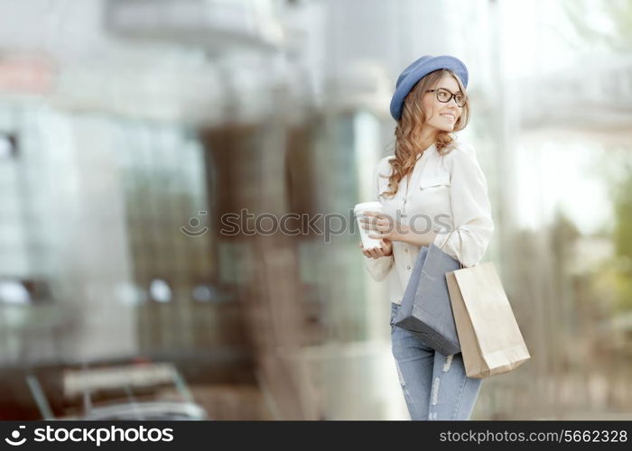 Happy young fashionable woman with bags having a coffee break after shopping and holding take away coffee against urban background.