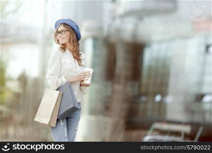 Happy young fashionable woman with bags having a coffee break after shopping and holding take away coffee against urban background.
