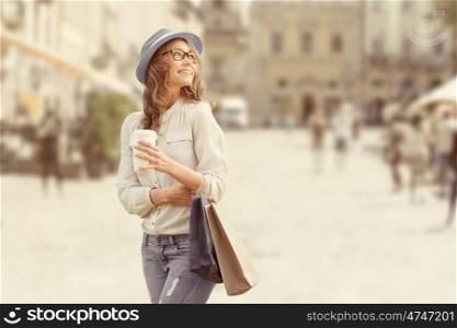 Happy young fashionable woman with shopping bags enjoying drinking coffee after shopping and holding take away coffee against urban background.