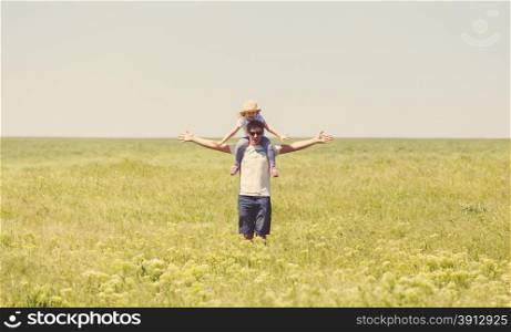 Happy young father holding his little daughter on shoulders against summer meadow