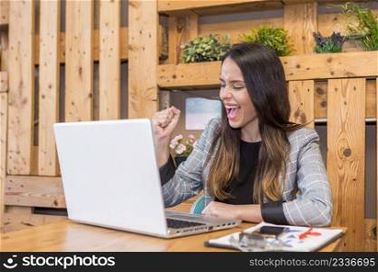 Happy young female freelancer clenching fist and yelling loud while celebrating successful achievement during remote work on laptop in cafe. Satisfied woman screaming with opened mouth while working online