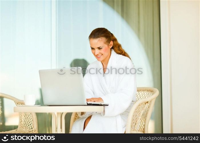 Happy young female in bathrobe sitting at table on terrace and using laptop