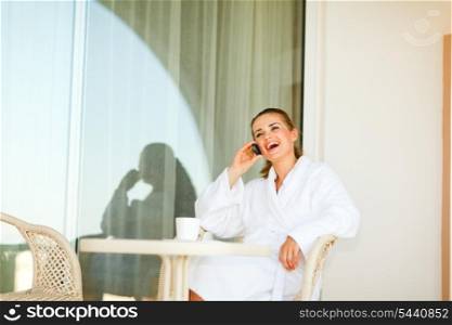 Happy young female in bathrobe speaking mobile phone on terrace