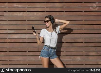 Happy young female in casual outfit and sunglasses smiling and browsing smart phone while listening to music and touching hair against timber wall. Smiling lady listening to music and using smartphone