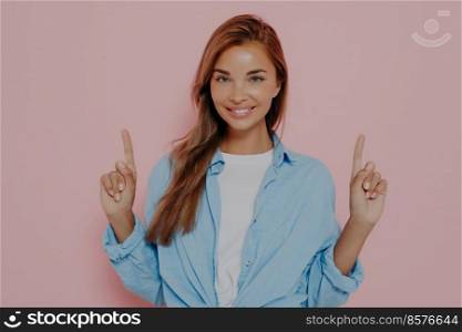 Happy young female points upwards with index fingers with broadly smile, shows awesome item on sale, wears white tshirt with blue shirt, isolated on pink studio wall. Promotion and advertisement. Happy young female points upwards with index fingers and smiles