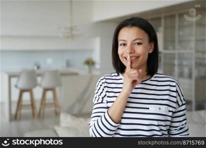 Happy young female puts index finger on lips, makes silence gesture, standing at home. Smiling woman asks to be quiet, to keep secret, looking at camera. Online store offer advertising.. Happy female makes silence gesture, asks to be quiet, to keep secret, looking at camera at home