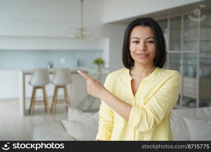 Happy young female satisfied client pointing finger aside, advertising good product or service. Friendly woman tenant homeowner makes interesting offer proposal, looking at camera, standing at home.. Female satisfied client pointing finger aside, advertising good product or service, standing at home