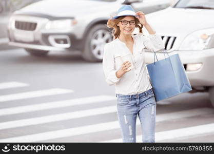 Happy young female student crossing the street with a coffee-to-go cup and tipping hat against urban city background.