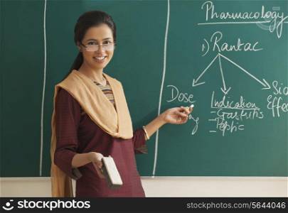 Happy young female teacher holding duster and chalk against green board