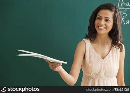 Happy young female teacher holding file while looking away against green board