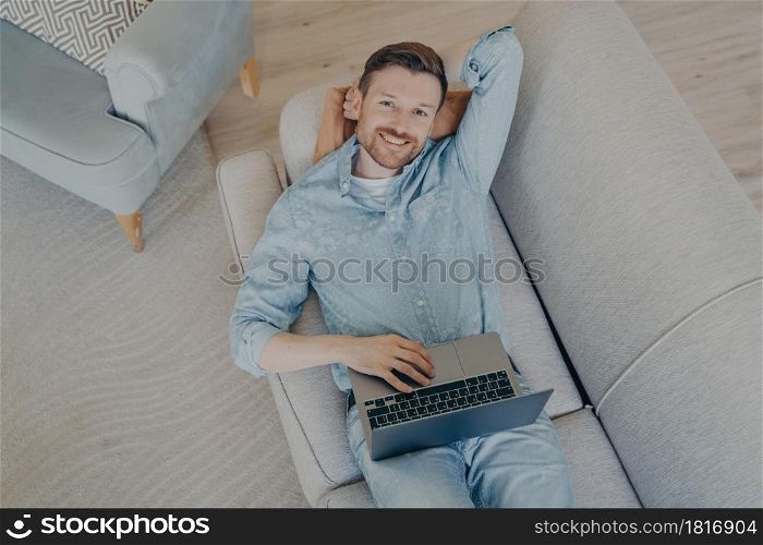 Happy young freelancer relaxing after finishing commission early, lying on comfortable couch in living room, resting head on hand, looking up while smiling and being satisfied. Happy young freelancer relaxing after finishing commission