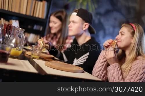 Happy young friends eating sandwiches and smiling while spending time together in cafe. Foreground attractive girl eating delicious sandwich with blurry mates enjoying meal on the background. Angle view.