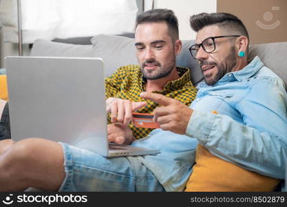 Happy young gay couple using laptop computer while sitting on a couch at home, shopping online with a credit card. High quality photography. Happy young gay couple using laptop computer while sitting on a couch at home, shopping online with a credit card