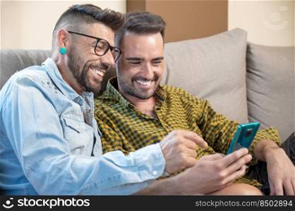 Happy young gay couple using mobile phone while sitting on a sofa in the living room. High quality photography.. Happy young gay couple using mobile phone while sitting on a sofa in the living room