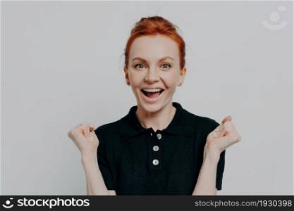 Happy young ginger girl in black polo t-shirt being excited by great news, keeping mouth open and raising hands with clenched fists, celebrating triumph, isolated on grey studio background. Happy young ginger girl being excited by great news, raising hands with clenched fists