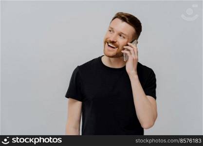 Happy young ginger man with stubble having pleasant conversation with friend or family, receiving positive news, guy with red hair looking aside and smiling joyfully, standing against grey studio wall. Happy man hearing good news on mobile phone