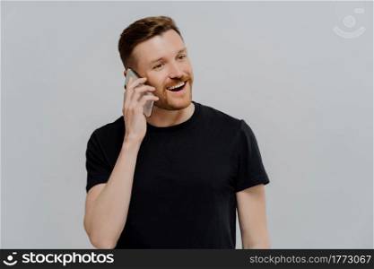 Happy young ginger man with stubble talking on mobile phone and feeling satisfied, receiving positive good news, guy with red hair looking aside and smiling while standing against grey studio wall. Excited man having pleasant conversation on mobile phone and feeling happy