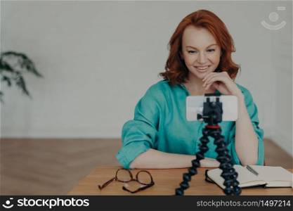 Happy young ginger woman shoots video in front of smartphone videocamera on tripod, gives recommendations how to start own business, has personal vlog, speaks on video call about online training