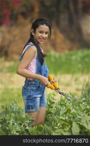 Happy young girl trimming plants in garden using a hedge shear or gardening scissors.