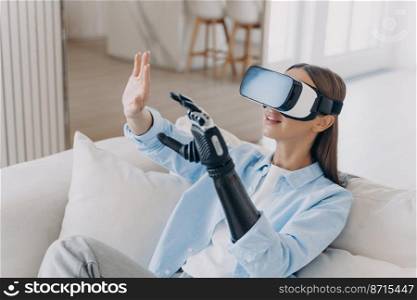 Happy young girl with disability in VR glasses touching objects of virtual reality by bionic prosthetic arm, sitting on sofa. Disabled woman interacts with cyberspace by artificial limb.. Girl with disability in VR glasses touching objects of virtual reality by bionic prosthetic arm