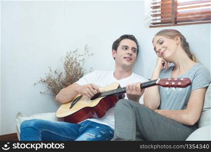 Happy young handsome caucasian man play guitar and sing song for his girlfriend sit in sofa in living room at apartment.Hipster couple having fun with play music together.