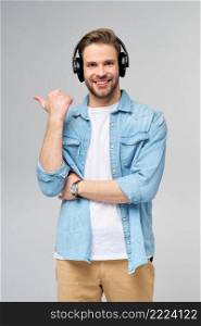 Happy young handsome man in jeans shirt pointing away standing against grey background wearing big headphones.. Happy young handsome man in jeans shirt pointing away standing against grey background wearing big headphones