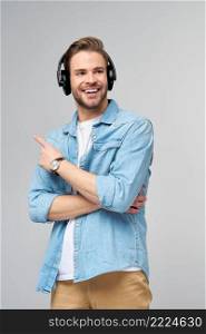 Happy young handsome man in jeans shirt pointing away standing against grey background wearing big headphones.. Happy young handsome man in jeans shirt pointing away standing against grey background wearing big headphones