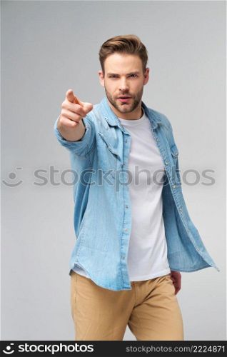 Happy young handsome man in jeans shirt pointing on camera standing against grey background.. Happy young handsome man in jeans shirt pointing on camera standing against grey background