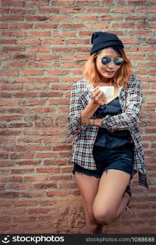 Happy young hipster woman standing against brick wall in town street with coffee cup in her hand.