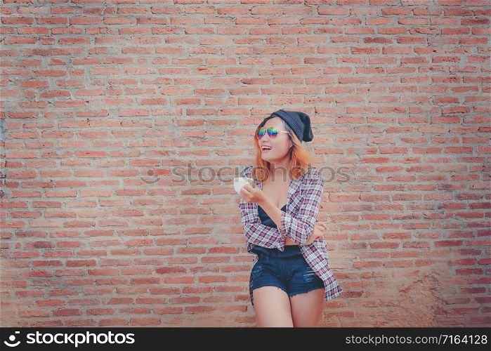 Happy young hipster woman standing against brick wall in town street with coffee cup in her hand.