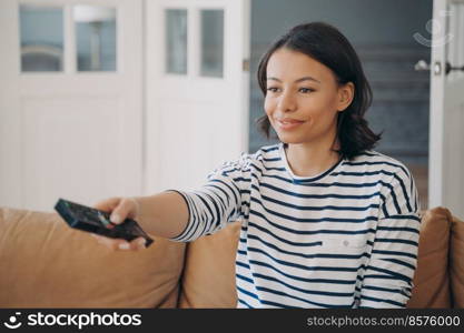 Happy young hispanic woman is shifting channels with remote control. Gorgeous lady is sitting on leather couch in cozy living room and smiling. Leisure and entertainment at home on quarantine.. Happy hispanic woman is shifting channels with remote control in cozy living room and smiling.