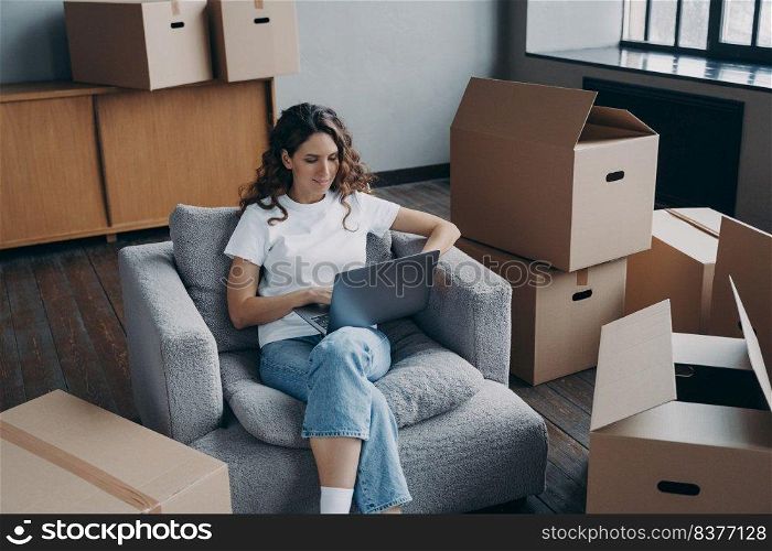 Happy young hispanic woman renting apartment online. Girl among cardboard boxes is using laptop and smiling. Happy homeowner is sitting in armchair with pc. Woman about to move.. Happy hispanic woman renting apartment online. Homeowner is sitting in armchair with pc.