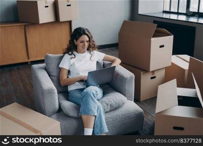 Happy young hispanic woman renting apartment online. Girl among cardboard boxes is using laptop and smiling. Happy homeowner is sitting in armchair with pc. Woman about to move.. Happy hispanic woman renting apartment online. Homeowner is sitting in armchair with pc.