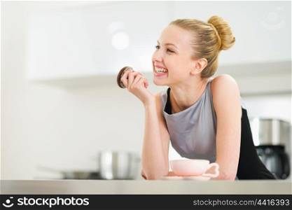 Happy young housewife drinking coffee with chocolate muffin in kitchen