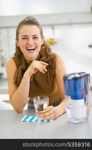 Happy young housewife drinking water from water filter pitcher in kitchen