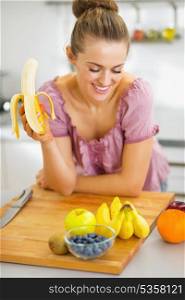 Happy young housewife eating banana in kitchen