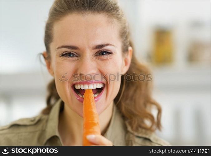 Happy young housewife eating carrot in kitchen