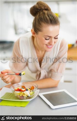 Happy young housewife eating fruits salad and using tablet pc