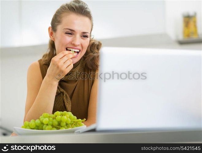 Happy young housewife eating grape and using laptop in kitchen
