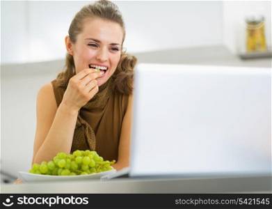 Happy young housewife eating grape and using laptop in kitchen