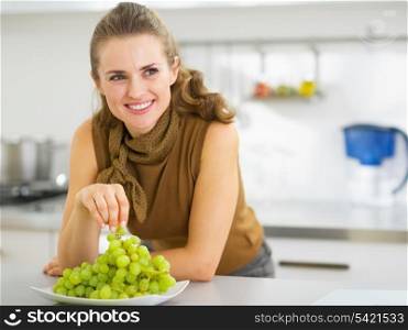 Happy young housewife eating grape in kitchen
