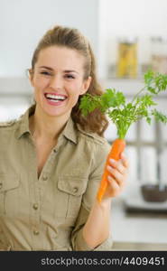 Happy young housewife holding carrot in kitchen