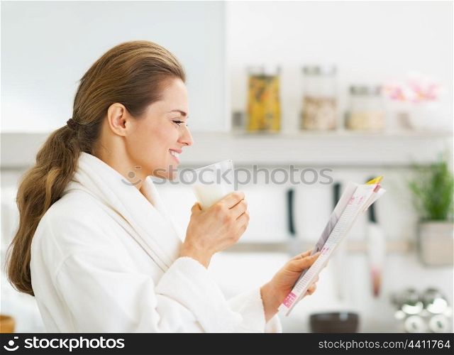 Happy young housewife in bathrobe drinking milk and reading magazine