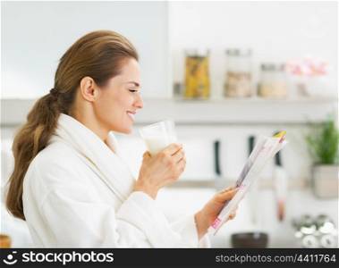 Happy young housewife in bathrobe drinking milk and reading magazine