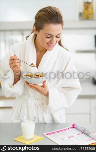 Happy young housewife in bathrobe having healthy breakfast and reading magazine