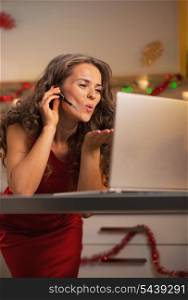 Happy young housewife in red dress having video chat on laptop in christmas decorated kitchen